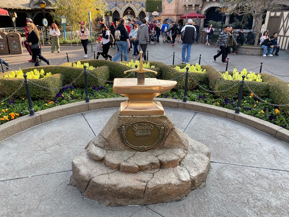 A golden sword stuck in an anvil with a plaque that reads 'The Sword in the Stone' in front of a castle.